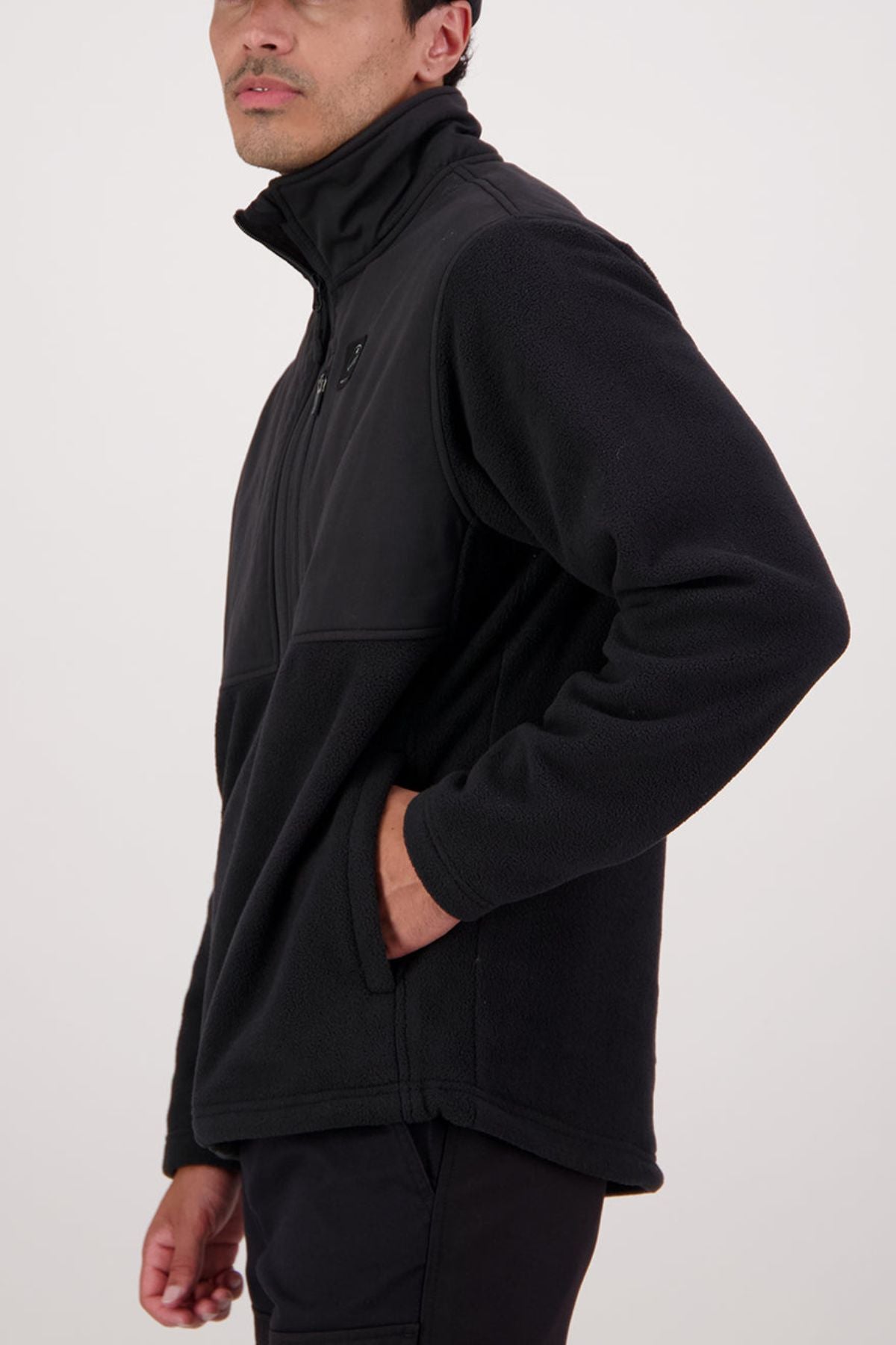 Someone wearing a Swanndri High Rock Hybrid Fleece Pullover in black, made from recycled polyester, stands sideways with one hand in a pocket against a plain light background.