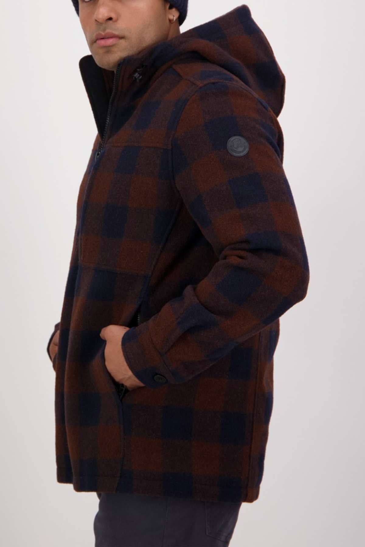 A person wearing a Swanndri Hudson Hoodie in brown and navy blue check stands sideways with one hand in their pocket against a plain background.