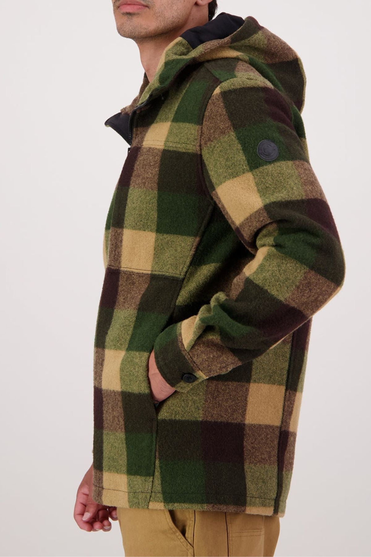 A person stands sideways against a plain background, left hand in the pocket of a green, brown, and tan plaid Swanndri Hudson Hoodie made from New Zealand wool.