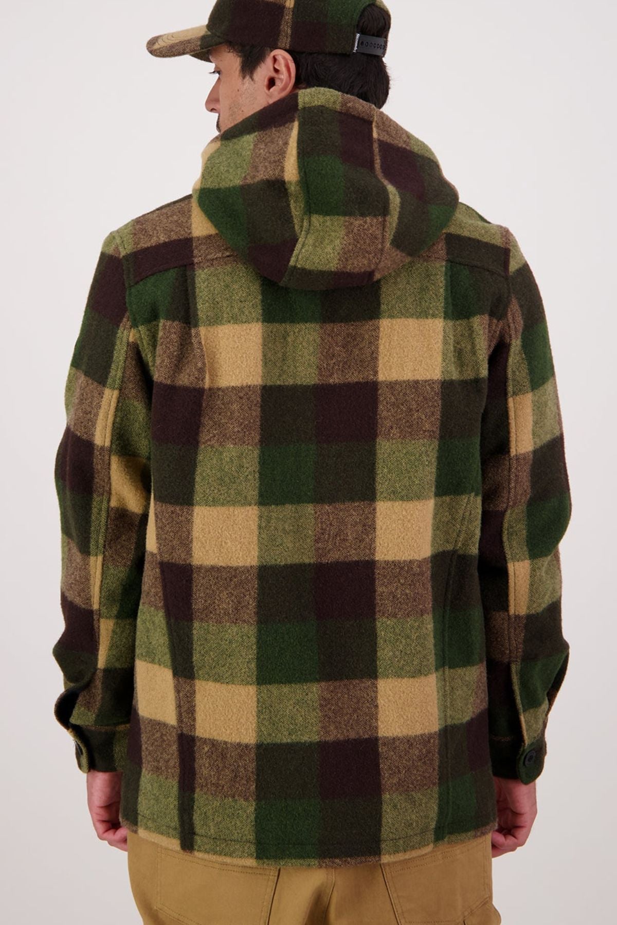 Facing away, a person wears the Swanndri Hudson Hoodie—a green, brown, and beige plaid New Zealand wool jacket—and a matching cap with tan pants, standing out against a plain light background.