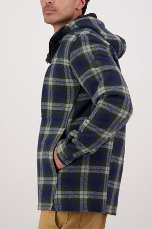 A man stands sideways with his hands in the pockets of a blue, green, and white plaid Swanndri Hudson Hoodie, a warm, durable New Zealand wool jacket featuring button-cuffed sleeves and a round sleeve patch.