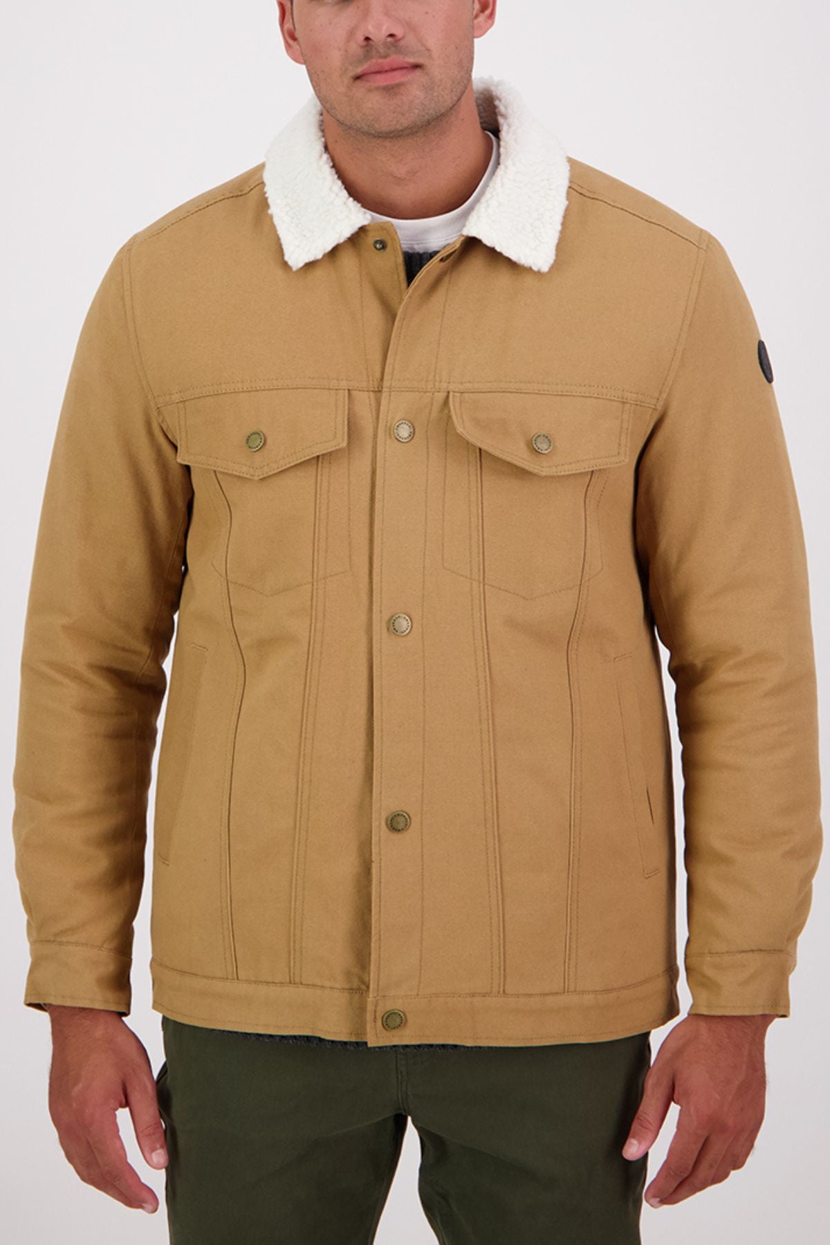 A man wears a Swanndri Kaituna Canvas Jacket with white sherpa lining, buttoned front, and two chest pockets, paired with green pants. The background is light and plain.
