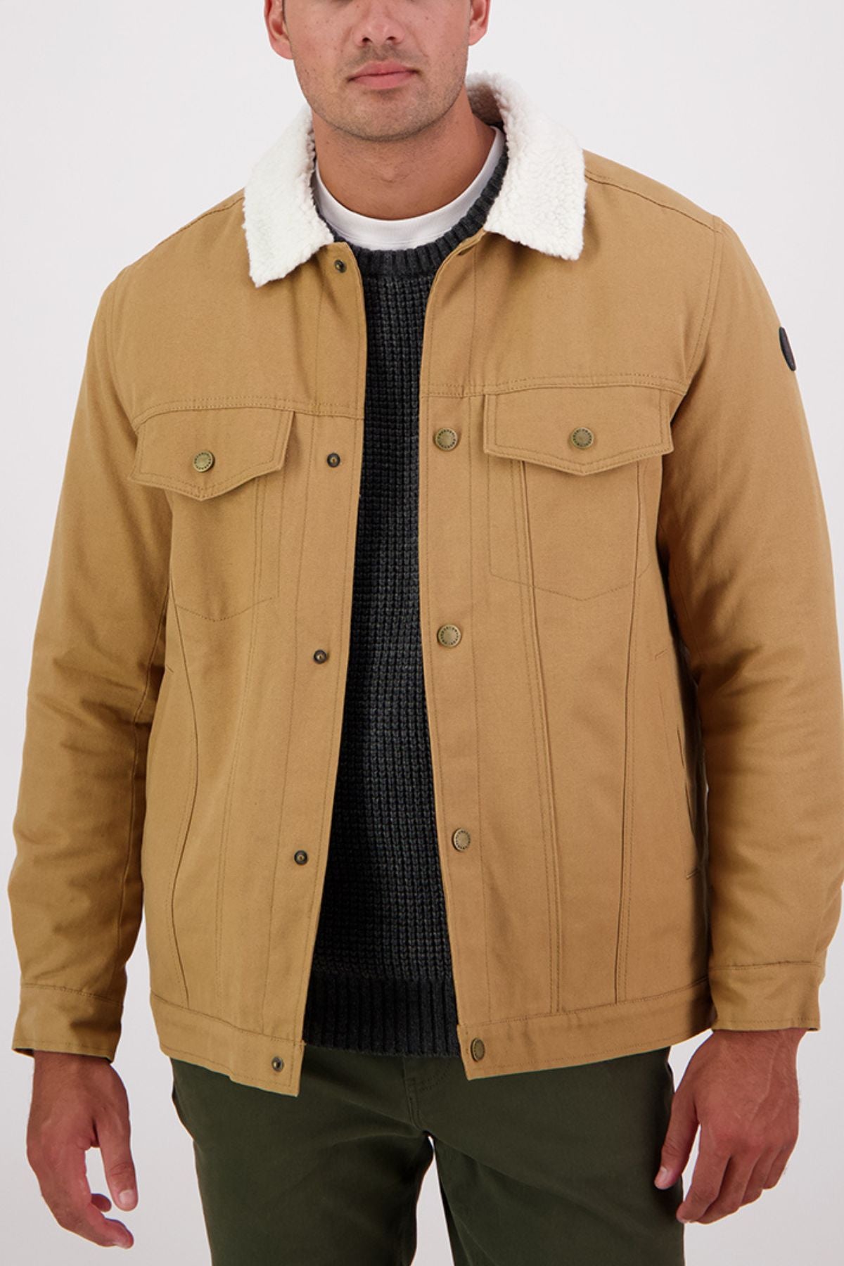 A man wears the Swanndri Kaituna Canvas Jacket with sherpa lining over a dark sweater and olive green pants, standing against a plain light background.