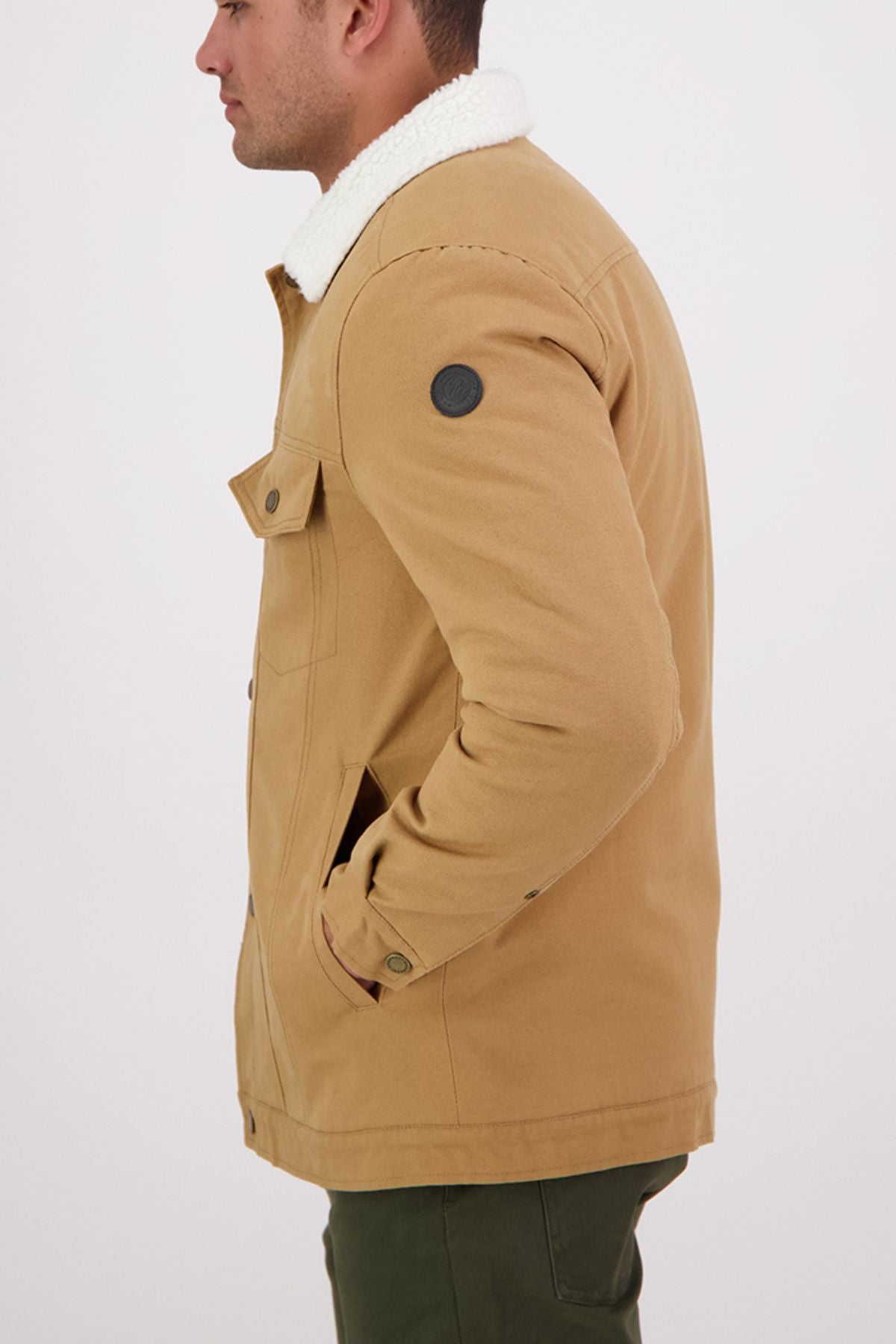 A person in side profile wears the Swanndri Kaituna Canvas Jacket—a tan, button-up jacket with sherpa lining, pockets, and a black circular sleeve patch—paired with green pants.