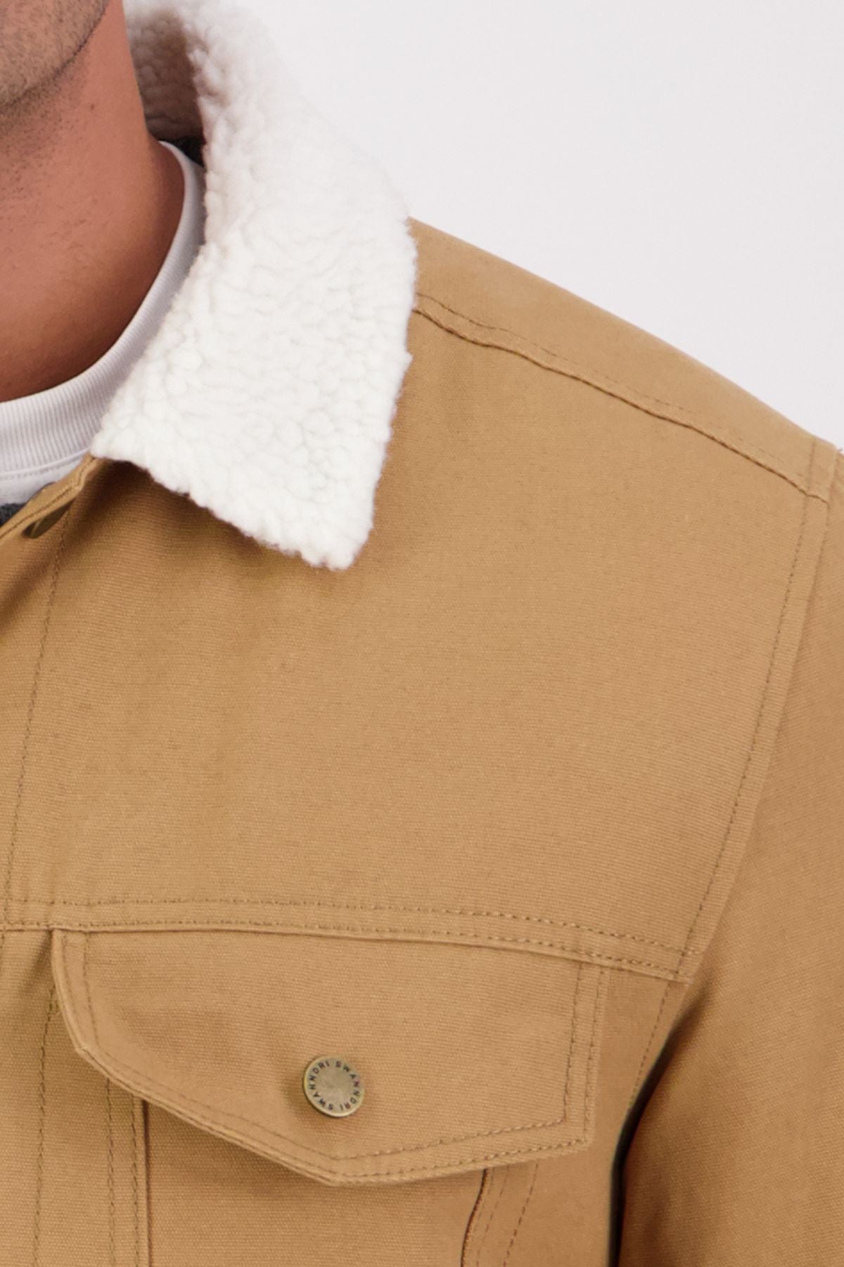 Close-up of the Swanndri Kaituna Canvas Jacket, featuring a white sherpa collar, brass chest pocket button, and a white shirt collar visible underneath. The image highlights the jacket’s upper left front detail.