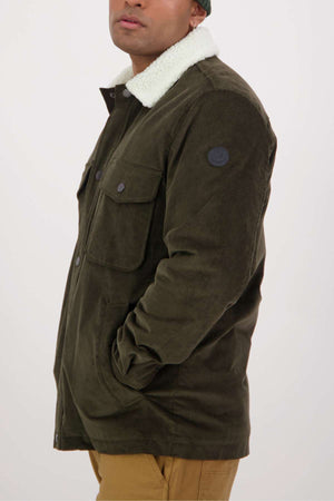 A person is shown from the side in a Swanndri Kaituna Corduroy Jacket with a white fleece collar and brown pants. The jacket, featuring polyester sherpa lining, front pockets, and a circular sleeve patch, is seen against a plain background with their face partially visible.
