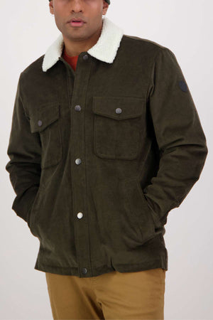 Wearing a Swanndri Kaituna Corduroy Jacket in dark green with a polyester sherpa collar and snap buttons, the person poses against a plain background. The jacket has two front pockets and is paired with a rust-colored shirt and tan pants.
