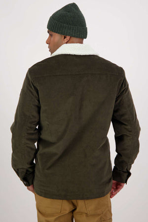 In a studio-lit setting against a plain background, a person stands with their back to the camera, wearing an olive Swanndri Kaituna Corduroy Jacket with polyester sherpa lining, paired with a green beanie and tan pants.