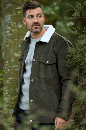 A bearded man with short hair wears the Swanndri Kaituna Corduroy Jacket, featuring a dark green color and white polyester sherpa lining, over a light gray sweater. He stands outdoors beside a tree amid lush greenery, gazing to the side.
