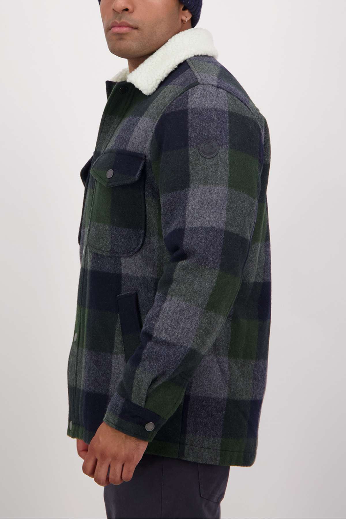 Wearing a Swanndri Kaituna Sherpa Lined Jacket in green and gray plaid with a warm sherpa lining and white collar, the person stands sideways, styled with a navy beanie and dark pants against a plain backdrop.