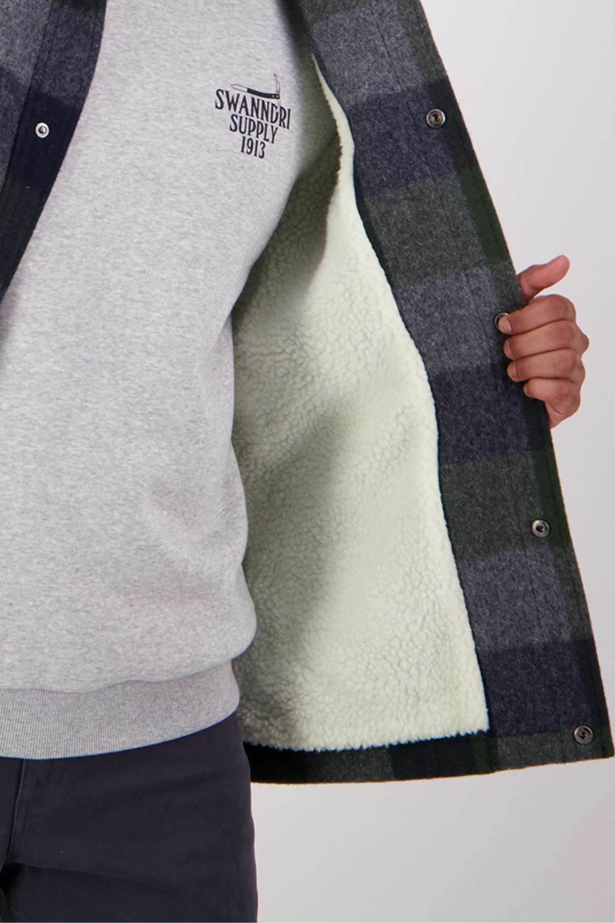 A person holds open a Swanndri Kaituna Sherpa Lined Jacket with green and gray plaid, revealing its cozy lining. They wear a gray sweatshirt with "Swandri Supply 1913" printed on it and dark pants.
