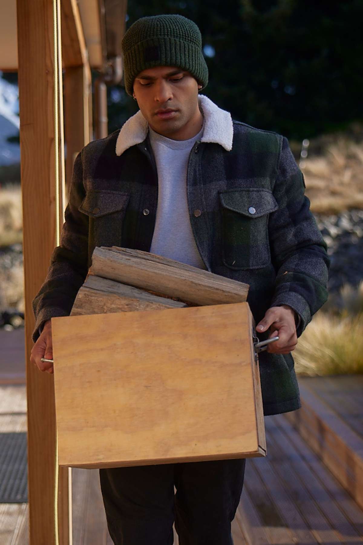 A person in a green beanie and Swanndri Kaituna Sherpa Lined Jacket carries a box of firewood on a wooden deck. Blurred greenery and part of a building in the background add warmth to the cozy scene.