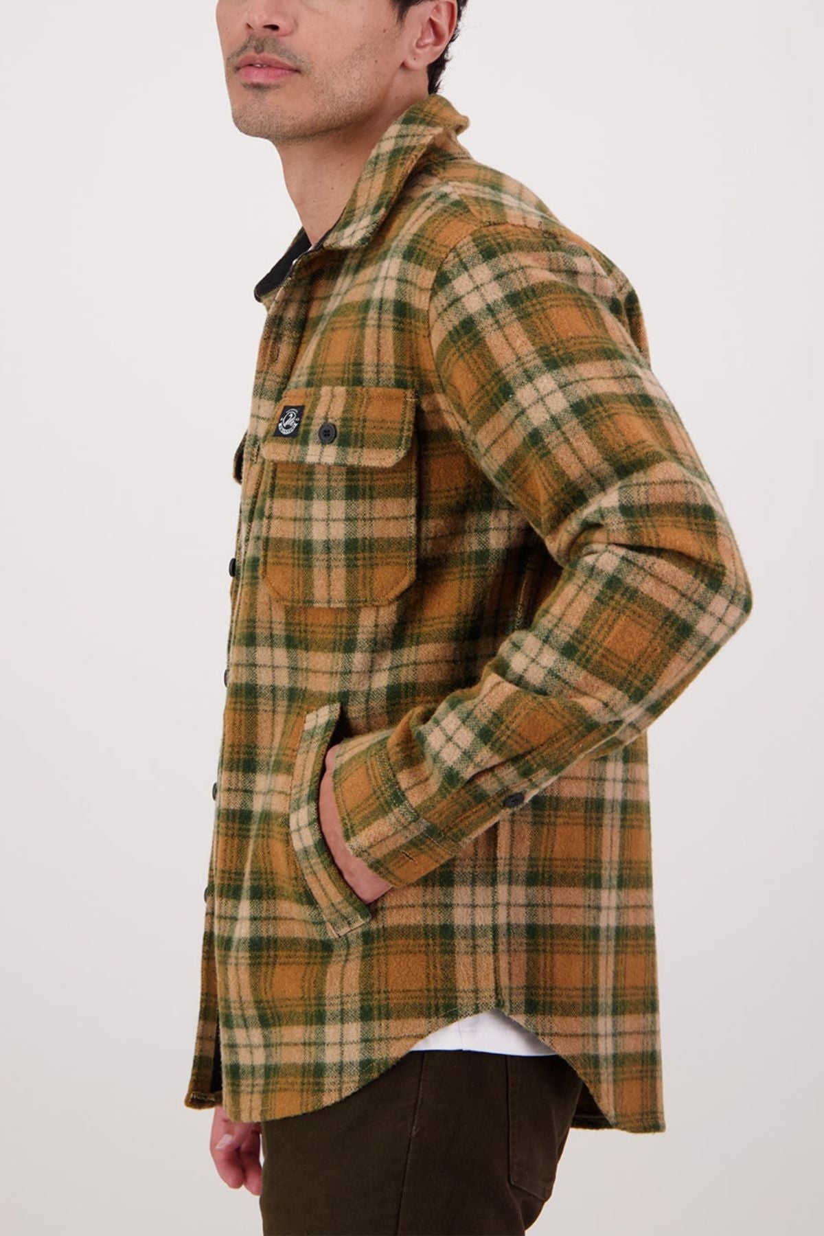 A man stands sideways with a hand in his pocket, wearing the Swanndri Kiaraki V4 Wool Shacket—a brown, tan, and green plaid button-up made from New Zealand wool with front and side pockets—against a plain light background.