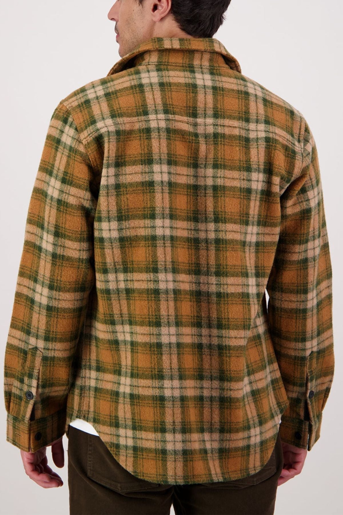 A person seen from behind wears the Swanndri Kiaraki V4 Wool Shacket—a brown, green, and beige plaid flannel shirt with heritage styling and dark buttons—standing against a plain light background.