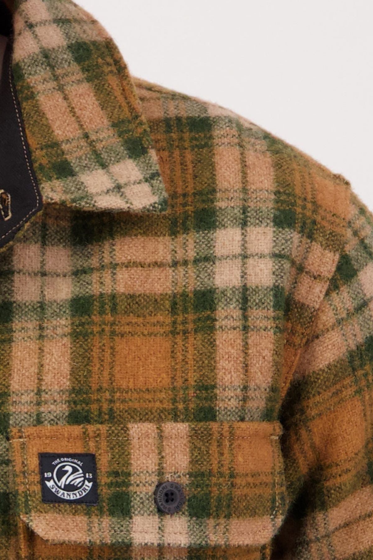 Close-up of the Swanndri Kiaraki V4 Wool Shacket—a green, tan, and cream plaid flannel shirt with a collar, black button, chest pocket featuring heritage styling, and a small black and white woven label.