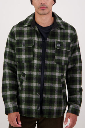 A man wearing a Swanndri Kiaraki V4 Wool Shacket in black and green plaid over a black t-shirt stands with relaxed hands by his sides against a plain light background.