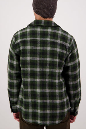 A person facing away from the camera wears a green and black plaid Swanndri Kiaraki V4 Wool Shacket with chest pockets, dark pants, and a knit beanie against a plain light background.