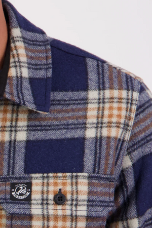 Close-up of the upper left section of the Swanndri Kiaraki v3 Wool Shacket, showing its blue, brown, and white plaid flannel pattern, buttoned chest pocket, and a small black-and-white brand logo on the pocket.