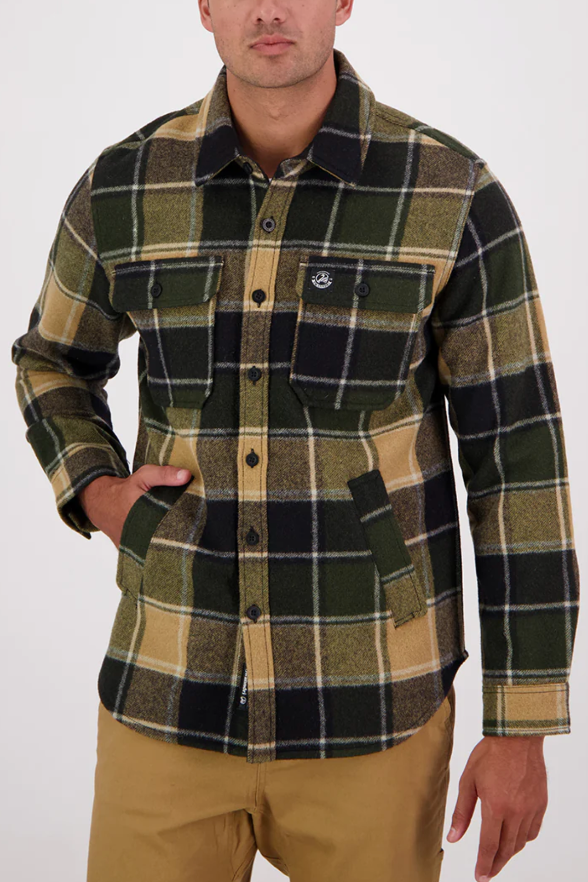 A man wearing a Swanndri Kiaraki v3 Wool Shacket in green, black, and tan plaid with two chest pockets and khaki pants stands against a plain light background, one hand in his pocket.