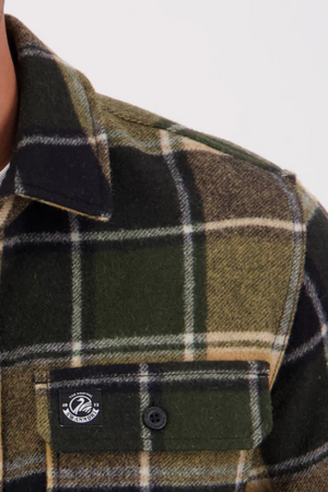 Close-up of the Swanndri Kiaraki v3 Wool Shacket in green, black, and beige plaid. Features a buttoned left chest pocket with a small round logo tag on the flap. The image focuses on the upper shoulder and chest area.