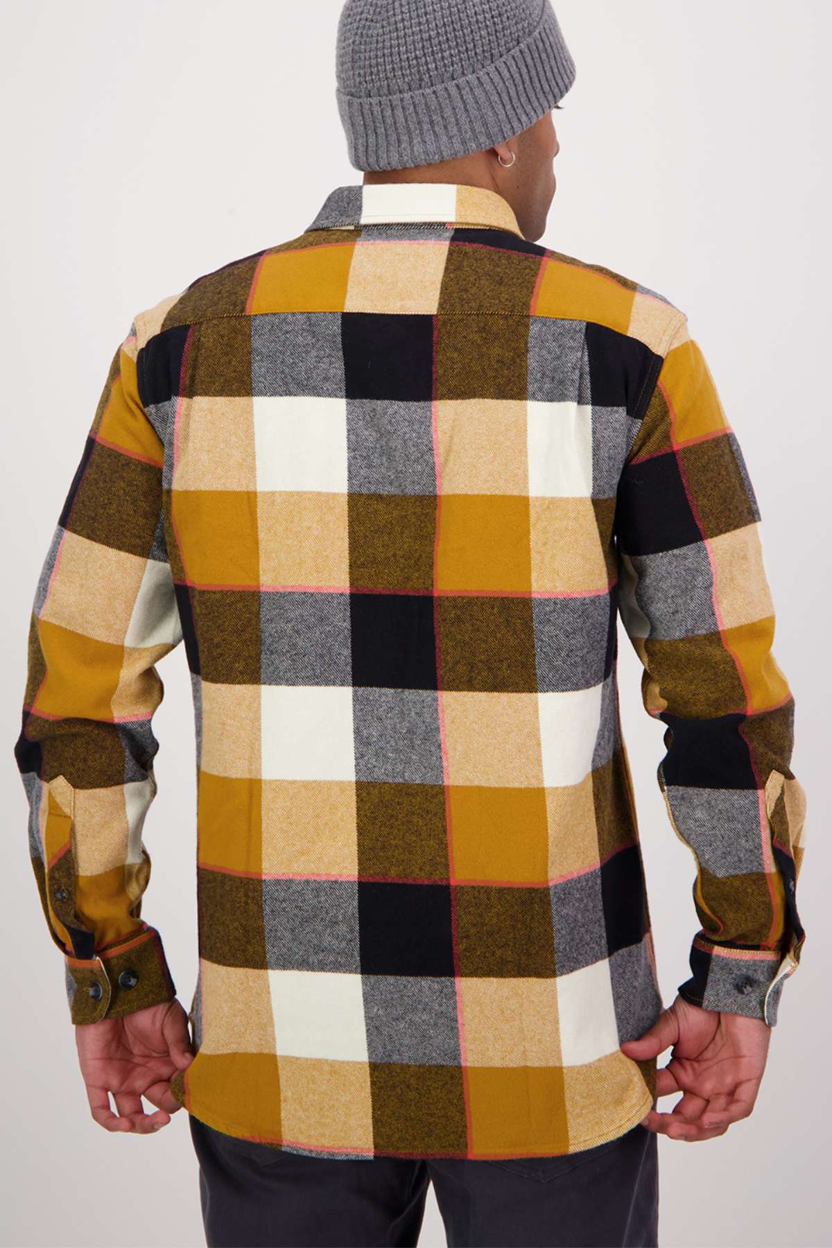 In a classic fit, checkered flannel shirt reminiscent of the Swanndri Lowcliffe Shacket, featuring 100% cotton in mustard, white, black, and gray shades. They pair it with dark pants and a gray beanie as they stand facing away from the camera.