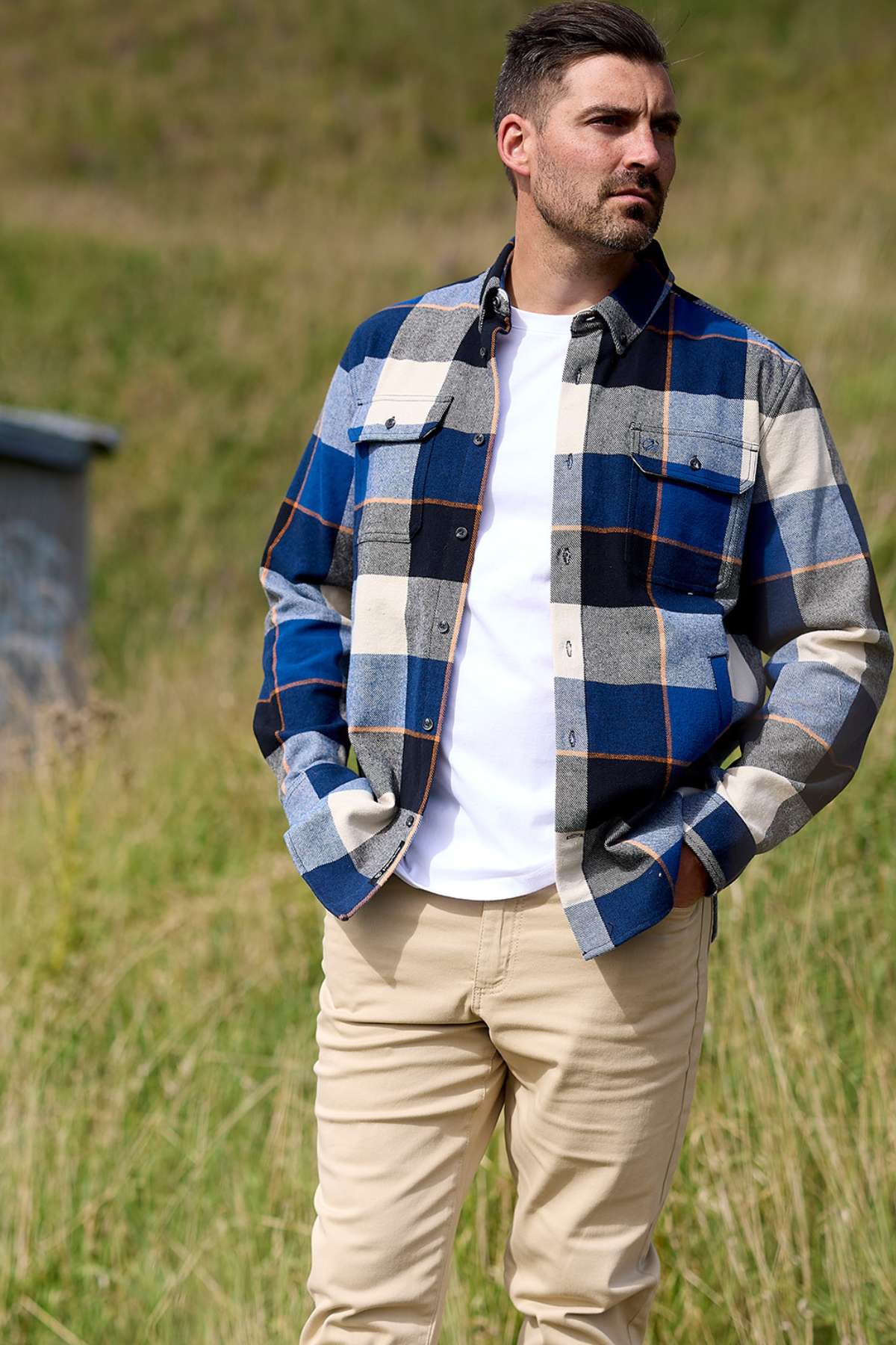 A man with short dark hair and a beard stands outside on grass, wearing a Swanndri Lowcliffe Shacket over a white T-shirt and beige pants, looking to the side with one hand in his pocket.