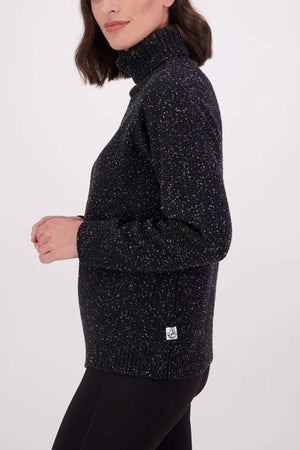 A woman with shoulder-length dark hair wears the Swanndri Lyttelton v2 Roll Neck Knit, a black speckled sweater, paired with black pants. She stands sideways with her left arm slightly bent against a plain white background.