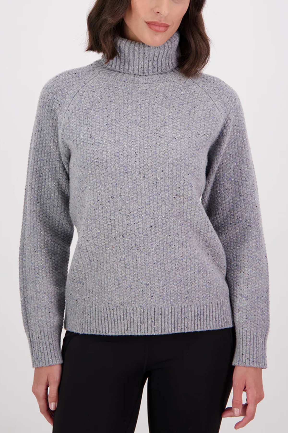 A woman in the Swanndri Lyttelton v2 Roll Neck Knit, a light gray ribbed jumper, and black pants stands against a plain background. Her shoulder-length wavy hair and the classic knit evoke timeless style.