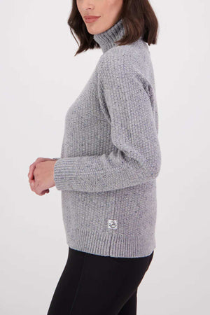 Woman in profile wears the Swanndri Lyttelton v2 Roll Neck Knit in light grey with black pants, standing against a plain white background. She has shoulder-length dark brown hair.