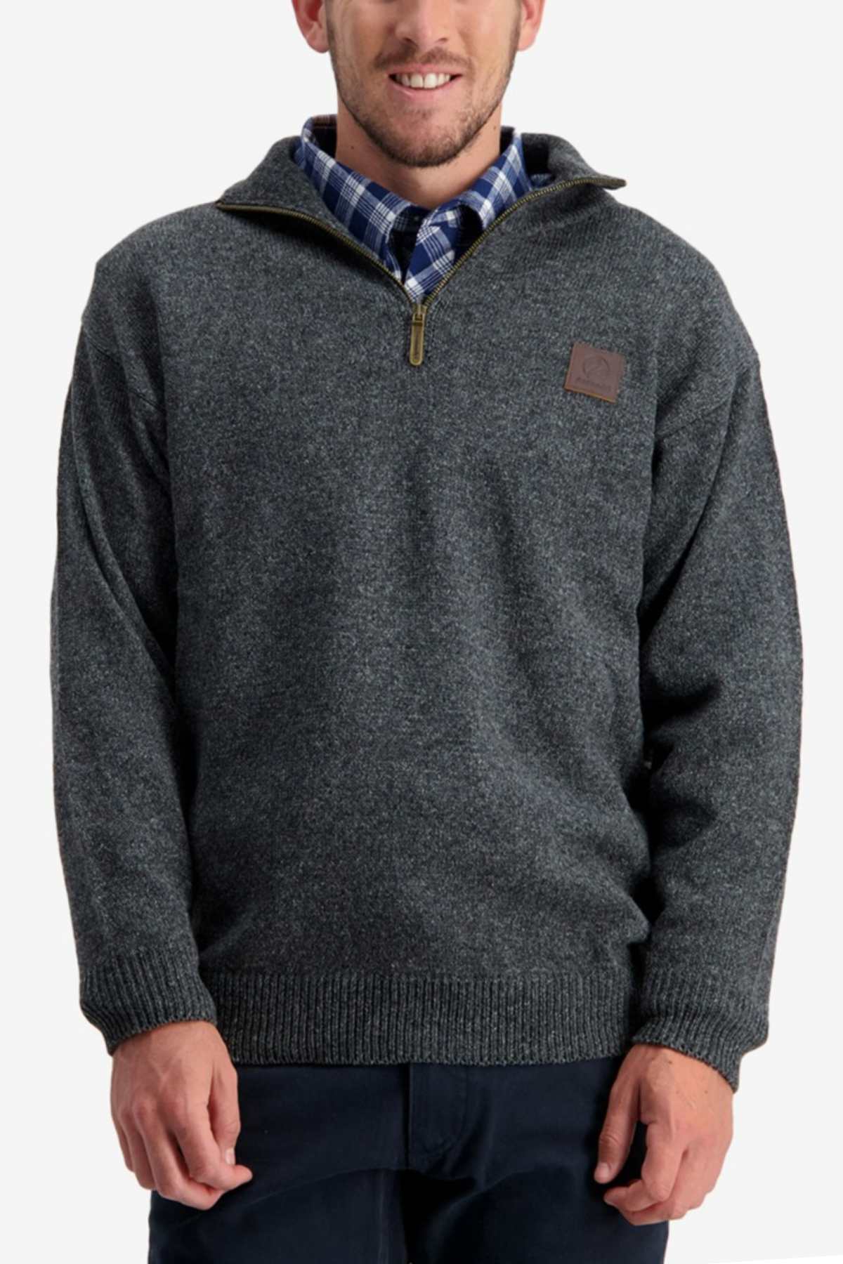 A man is wearing a Swanndri Mariner Zip Neck Sweater in dark gray over a blue and white plaid shirt. The knit sweater features a ribbed hem and cuffs, with a leather patch on the left chest. He is standing against a plain light background.