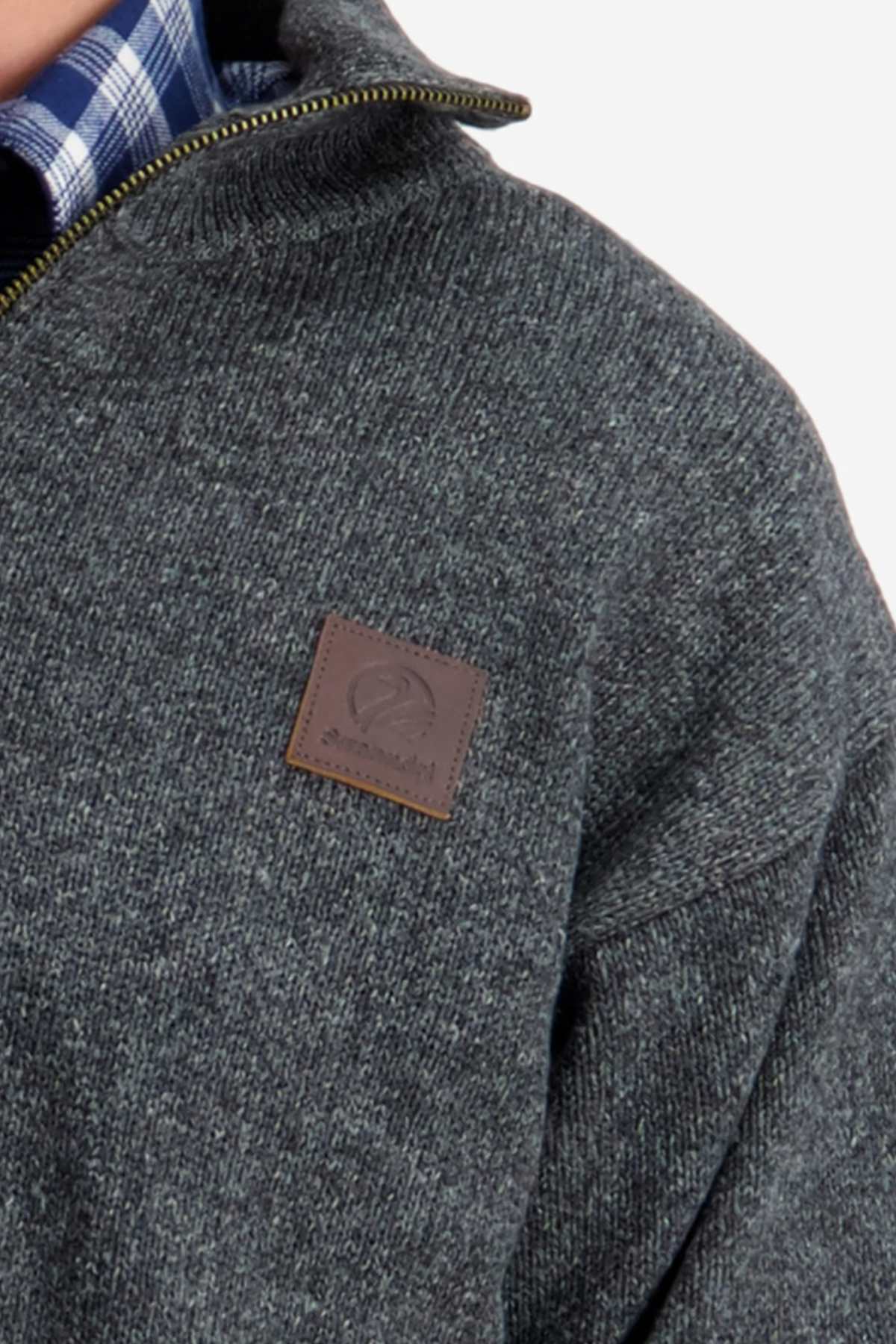 Close-up of a person wearing the Swanndri Mariner Zip Neck Sweater in dark gray with a half zip front and a visible leather patch logo on the chest. The collar of a blue and white plaid shirt peeks out from underneath the wool-polyester blend sweater. The background is plain and light-colored.