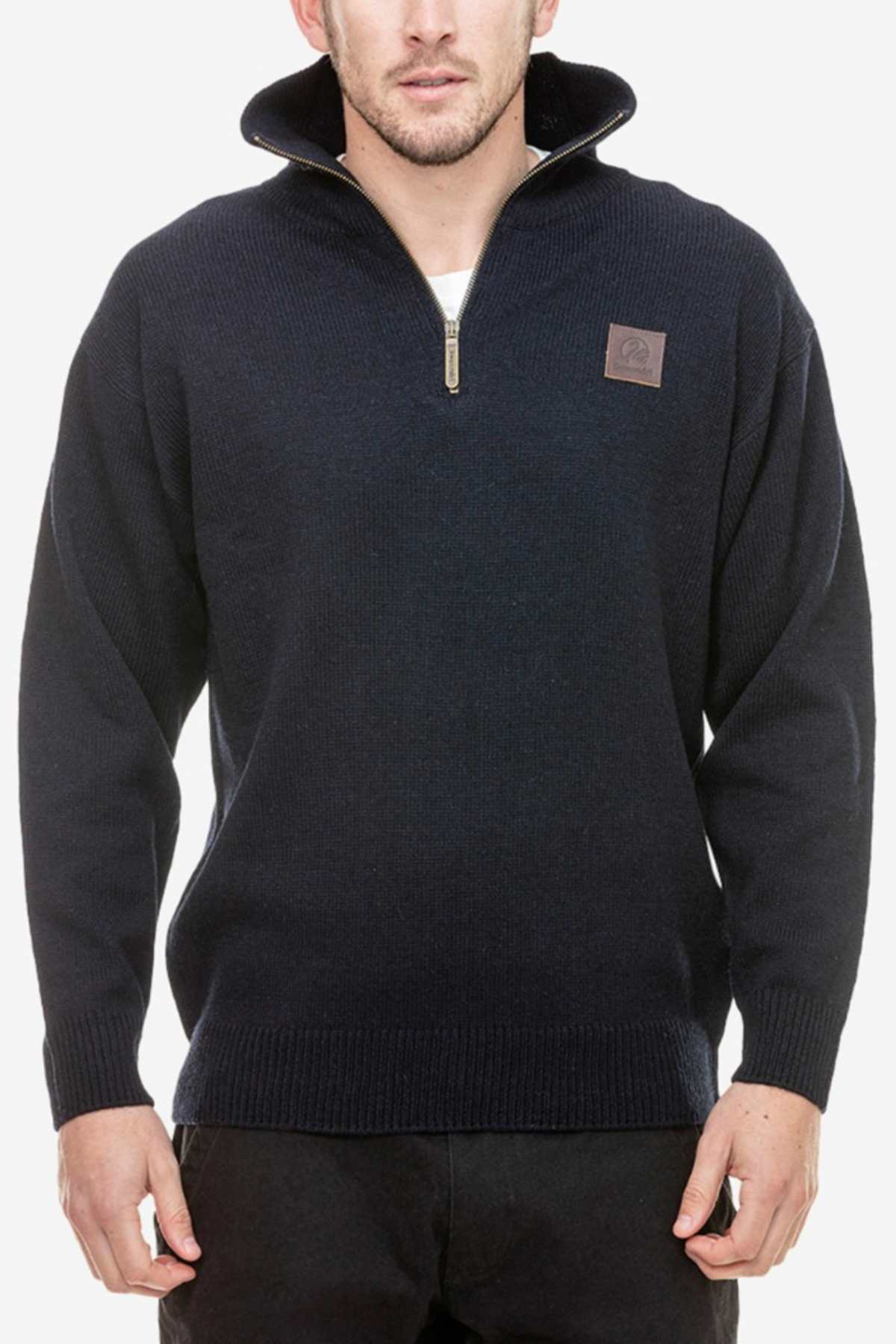 A man with short hair wears the Swanndri Mariner Zip Neck Sweater, a dark navy, long-sleeved wool knit featuring a half zip collar and a small leather patch on the left chest. He pairs it with black pants and stands against a white background.