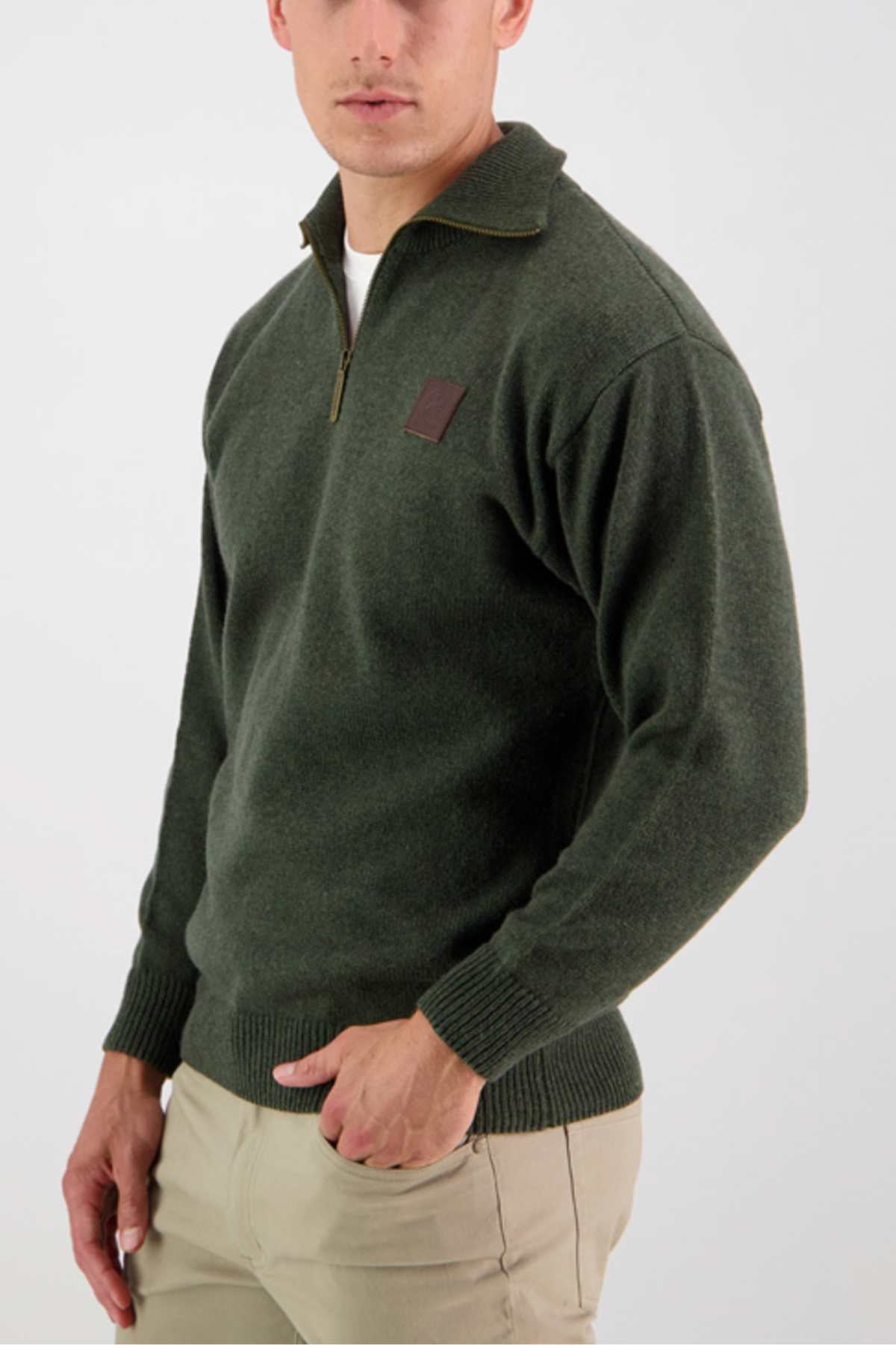 A person is wearing a Swanndri Mariner Zip Neck Sweater in dark green, featuring a brown patch on the chest, ribbed cuffs, and a ribbed hem, paired with beige pants. The person has their right hand in their pants pocket and is looking slightly to the left against a plain white background.