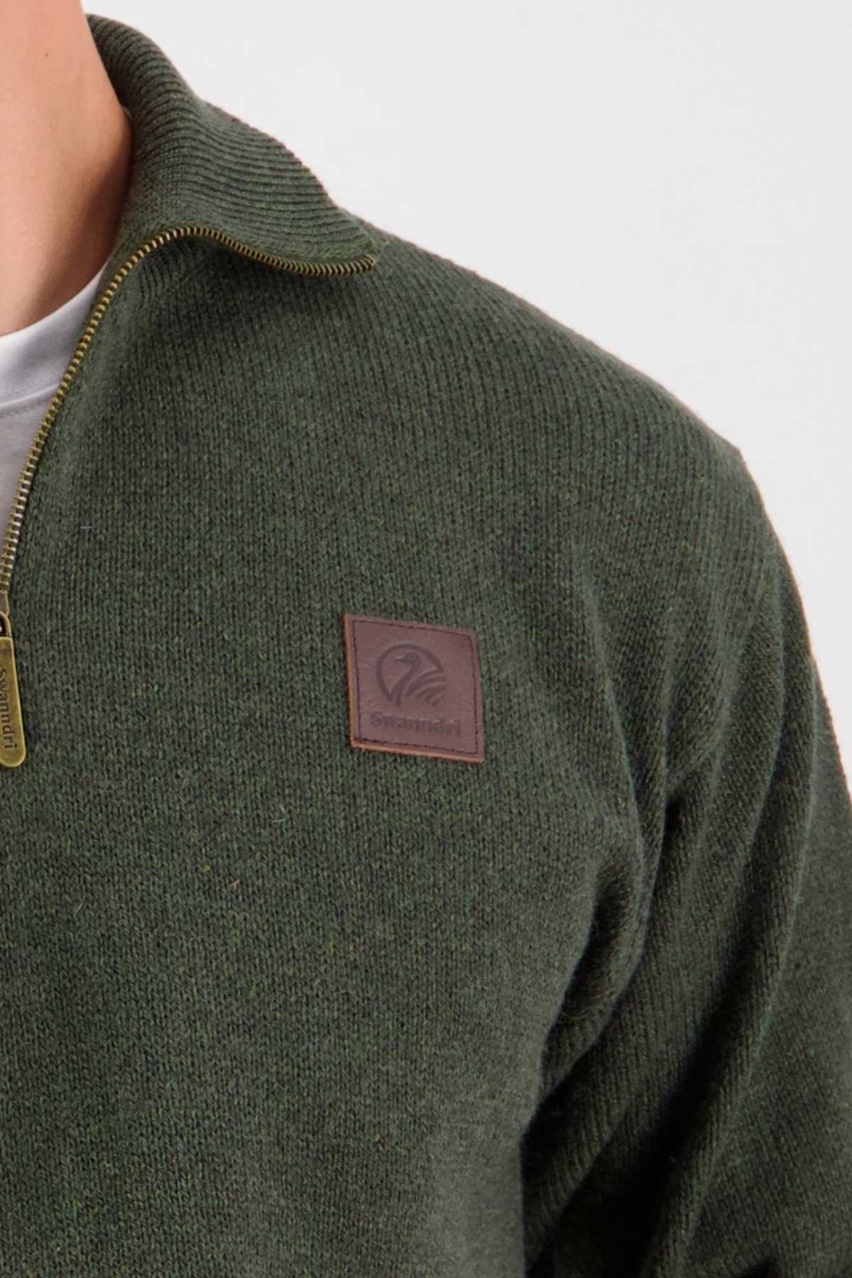Close-up of a person wearing the Swanndri Mariner Zip Neck Sweater in dark green wool blend knit, featuring a half zip front. The sweater has a brown rectangular leather patch on the chest embossed with the logo and is layered over a white shirt.
