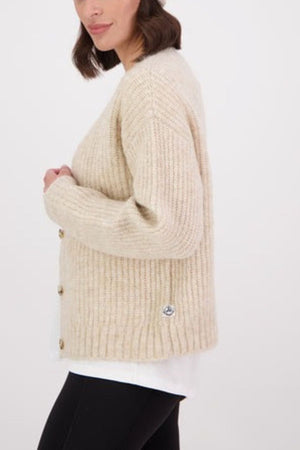 A woman stands sideways against a plain background, wearing the Swanndri Marydale Cardigan—a beige, chunky knit mohair wool blend—styled over a white top and black pants for a sustainable fashion look.