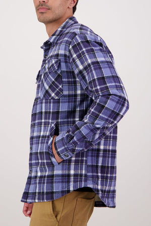 A person wearing a blue and purple plaid Swanndri Morven Sherpa Shirt with tan pants stands sideways, one hand in their pocket, the other by their side, against a plain light background.