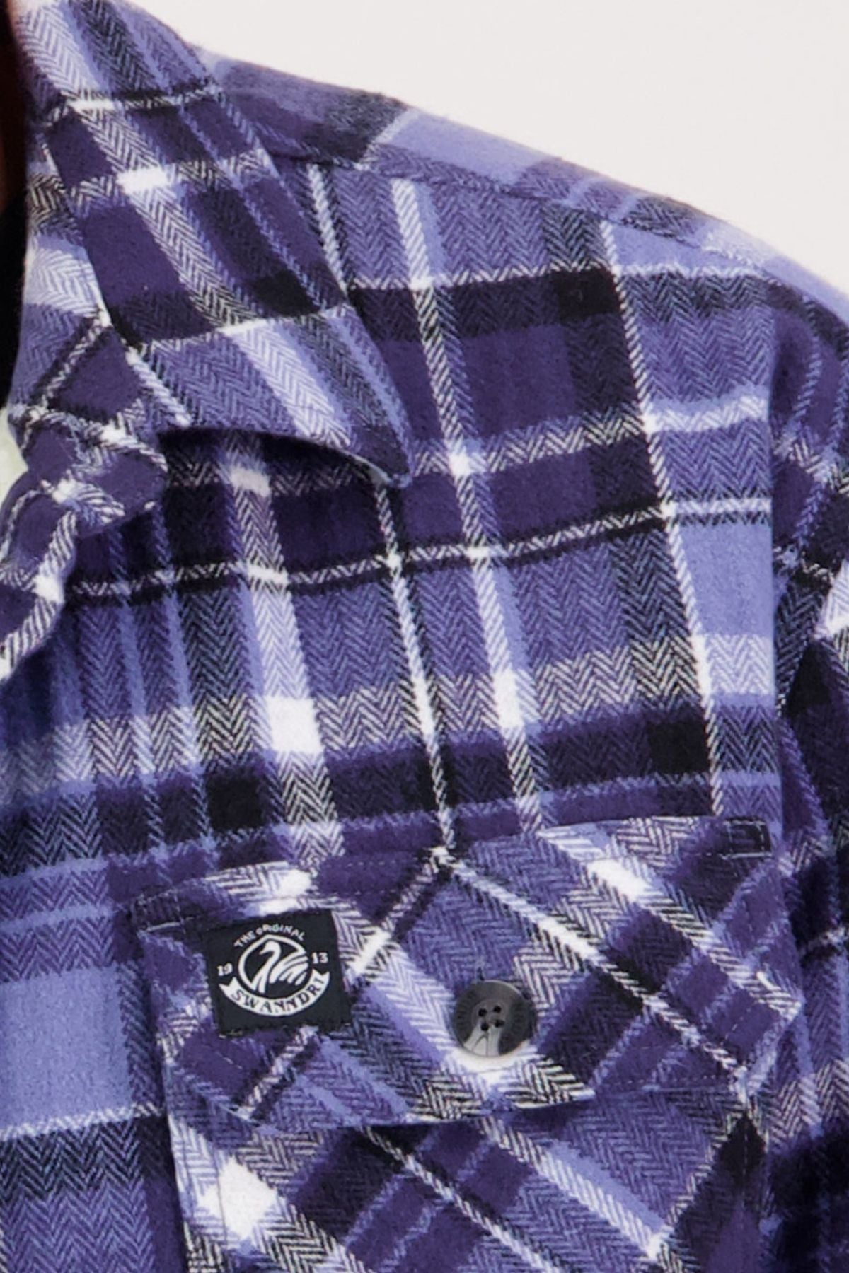 Close-up of the Swanndri Morven Sherpa Shirt in purple, black, and white plaid, featuring a buttoned breast pocket, a black and white logo tag above the flap, and warm ultra-soft sherpa fleece lining.