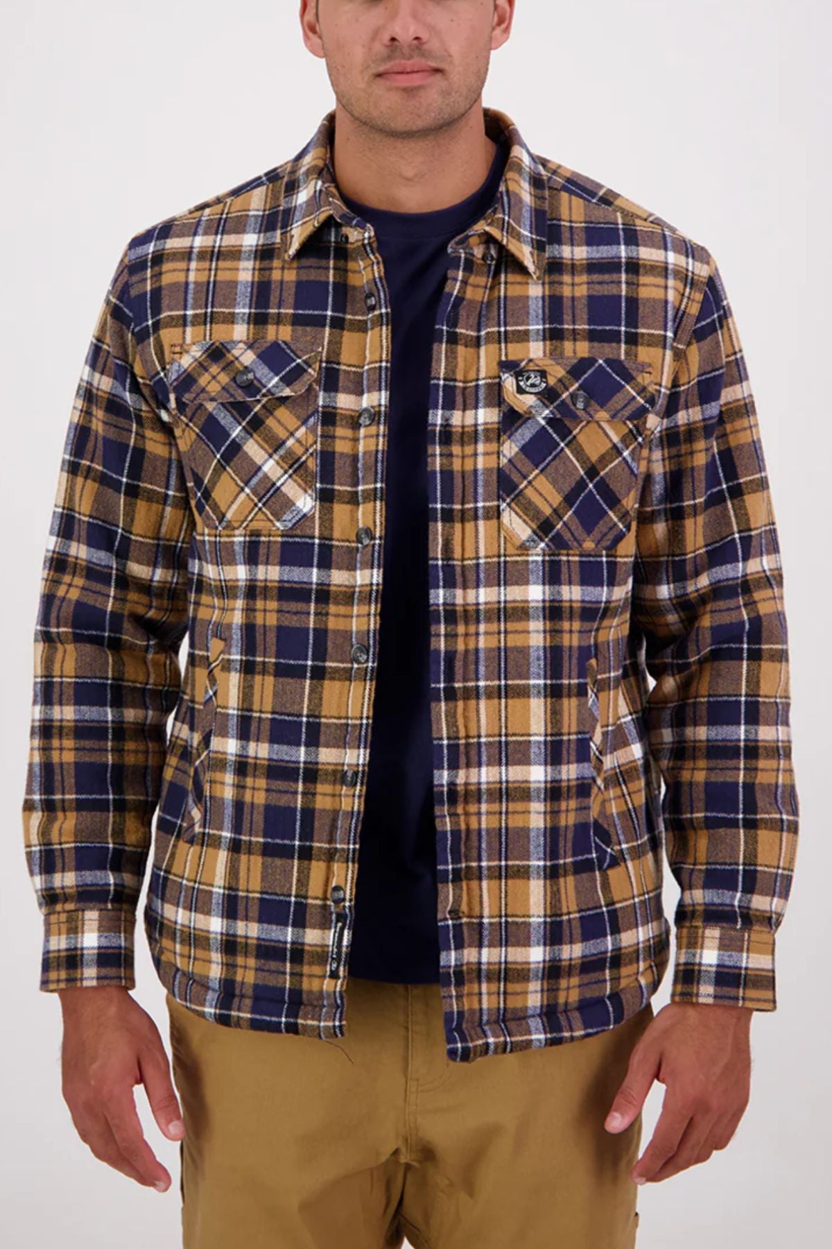 A man in a Swanndri Morven Sherpa Long Sleeve Shirt featuring blue, brown, and white 100% cotton fabric, layered over a dark navy t-shirt and khaki pants, stands against a plain light background.