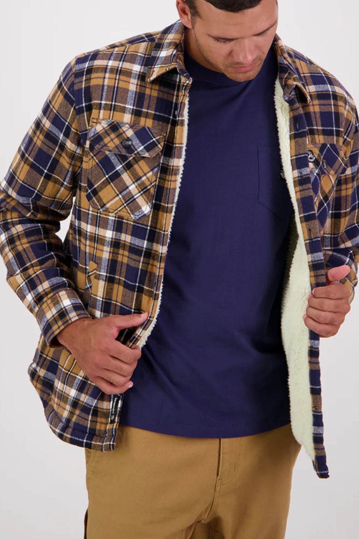A man wearing a Swanndri Morven Sherpa Long Sleeve Shirt—a blue and brown plaid, 100% cotton flannel with cosy sherpa fleece lining—over a navy t-shirt and tan pants looks down while opening the jacket.