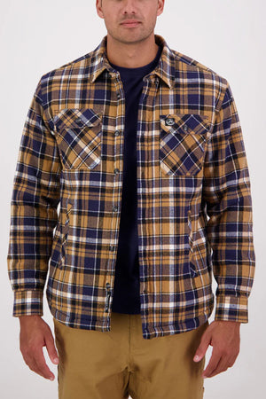 A man in a Swanndri Morven Sherpa Long Sleeve Shirt featuring blue, brown, and white 100% cotton fabric, layered over a dark navy t-shirt and khaki pants, stands against a plain light background.
