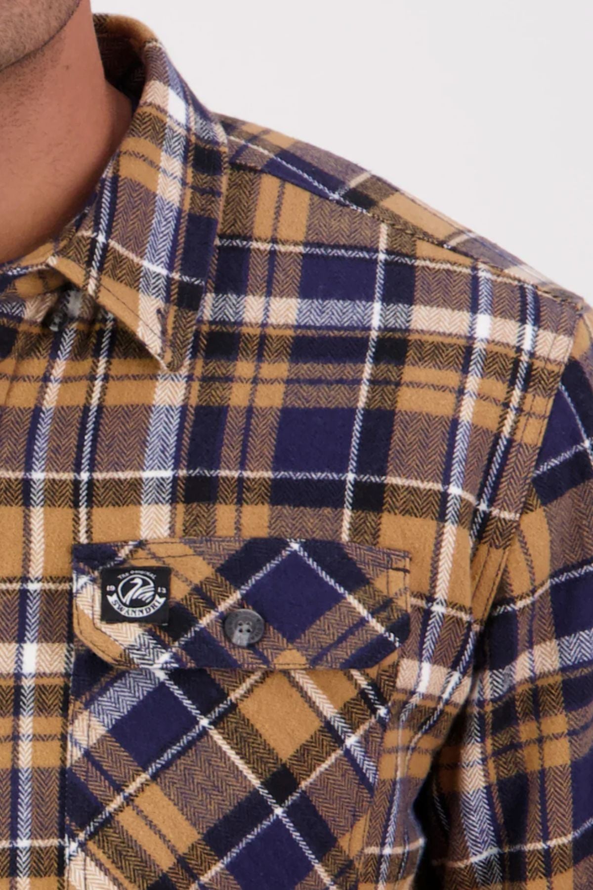 Close-up of a person wearing the Swanndri Morven Sherpa Long Sleeve Shirt in yellow, navy blue, and white plaid. The shirt features a buttoned chest pocket and black and white logo patch. Only the shoulder and chest are visible. Made from 100% cotton.