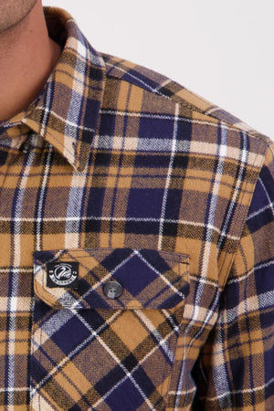 Close-up of a person wearing the Swanndri Morven Sherpa Long Sleeve Shirt in yellow, navy blue, and white plaid. The shirt features a buttoned chest pocket and black and white logo patch. Only the shoulder and chest are visible. Made from 100% cotton.