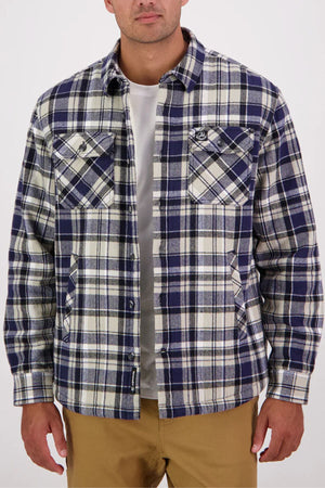 A man wearing a Swanndri Morven Sherpa Long Sleeve Shirt—blue, white, and gray plaid flannel over a white t-shirt and tan pants—stands against a plain light background.
