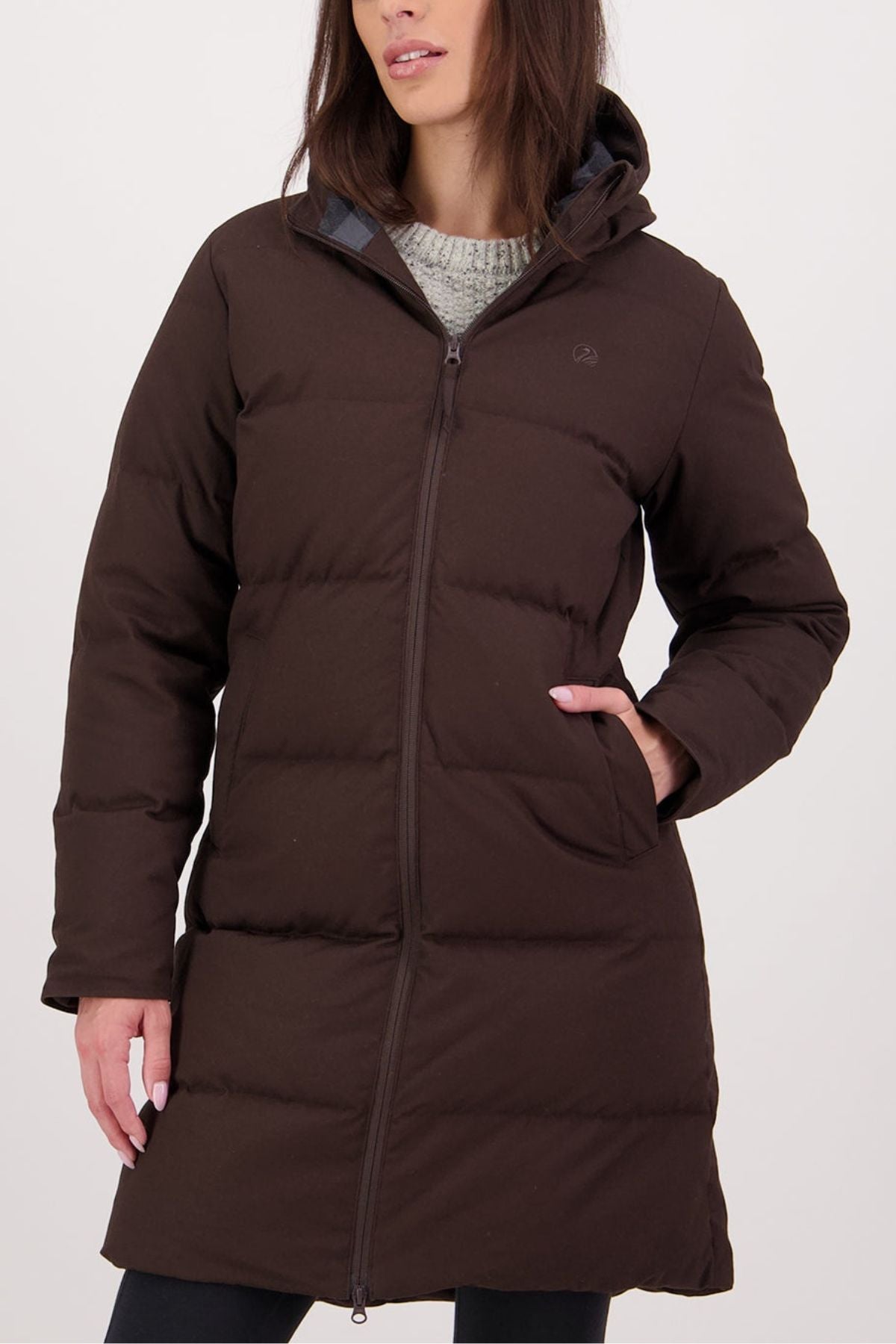 A woman stands against a plain light background, one hand in her pocket, wearing the Swanndri Nimbus Longline Down Coat—long, dark brown, featuring a hood and front zipper. Her face is partially visible.