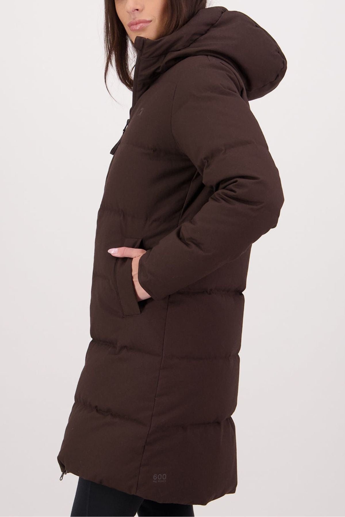 A woman stands in profile, hands in pockets, wearing the Swanndri Nimbus Longline Down Coat—an insulated long, dark brown hooded jacket—against a plain light background.