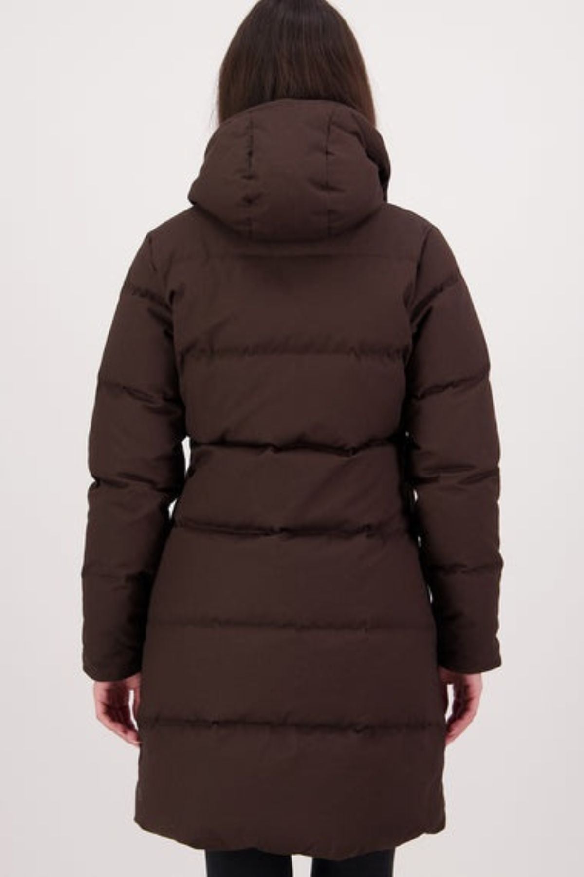 A person with long brown hair is seen from the back wearing a dark brown Swanndri Nimbus Longline Down Coat with a hood, horizontal quilting, and thigh-length fit against a plain light background.