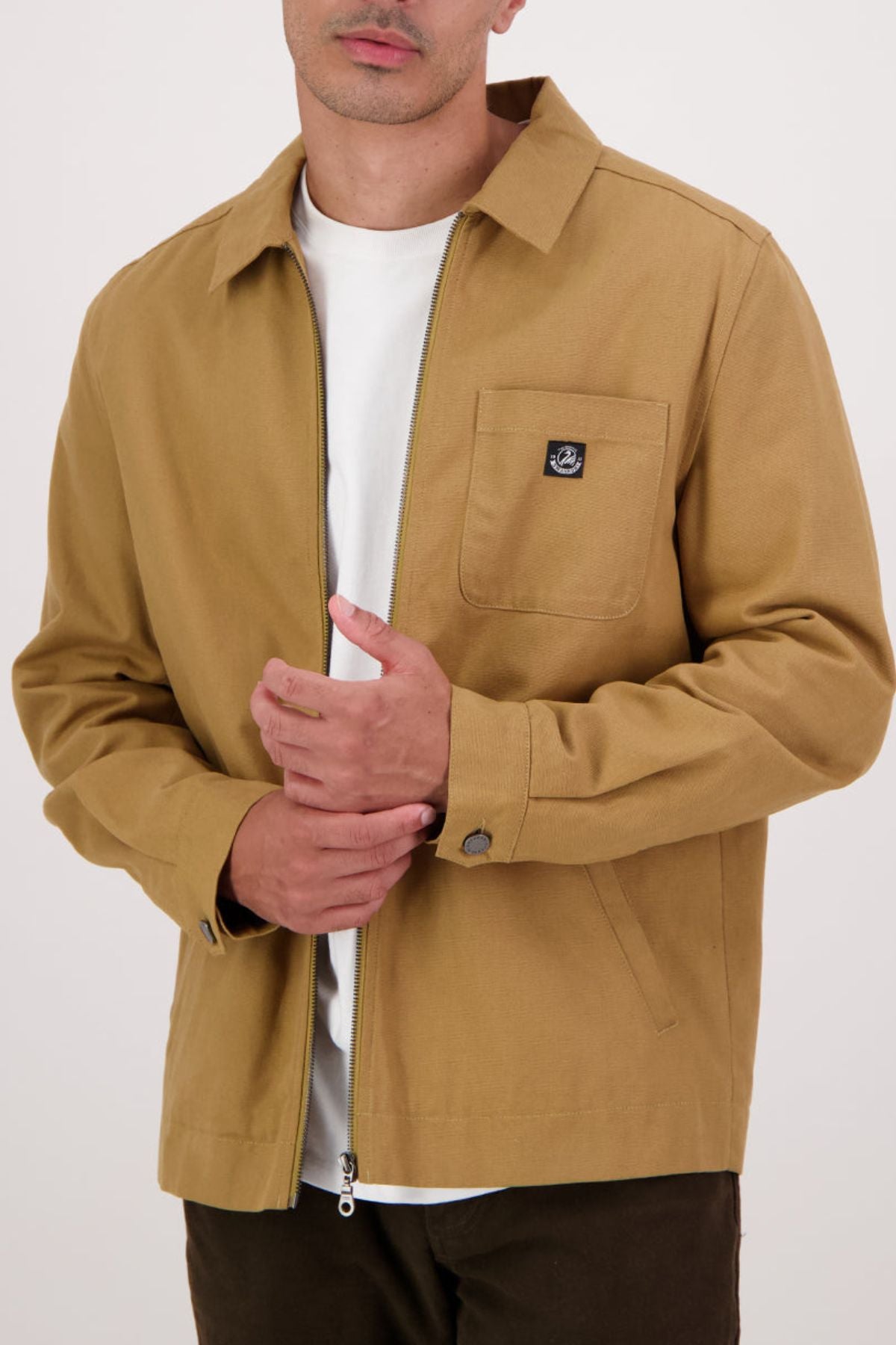 A person wears the Swanndri Northgate Canvas Jacket in tan over a white t-shirt, touching their chest. The jacket features a chest pocket with a small black logo patch and buttoned cuffs, set against a plain light background.