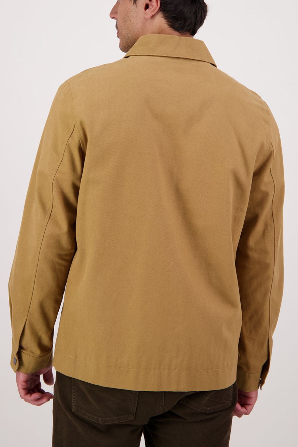 A man is shown from the back wearing a Swanndri Northgate Canvas Jacket in tan with long sleeves, a collar, and buttoned cuffs, paired with dark brown pants. The background is plain and light-colored.