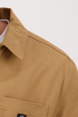 Close-up of a person wearing the Swanndri Northgate Canvas Jacket in tan; sturdy fabric, front pocket with stitched label, and only the shoulder, collar, and part of the chest are visible.