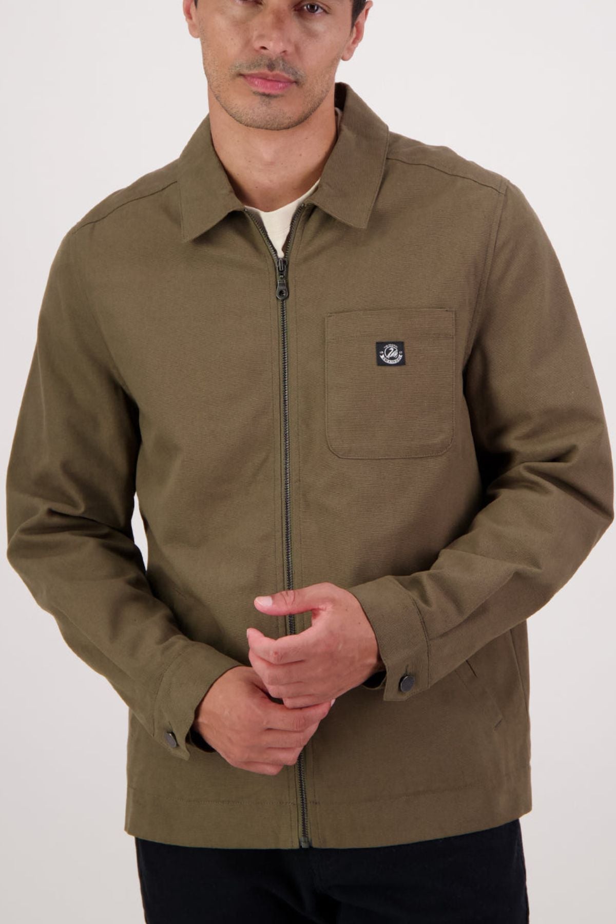 A man with short hair and a neutral expression stands against a plain light background, wearing the Swanndri Northgate Canvas Jacket in olive green, featuring a chest pocket and a small black logo.
